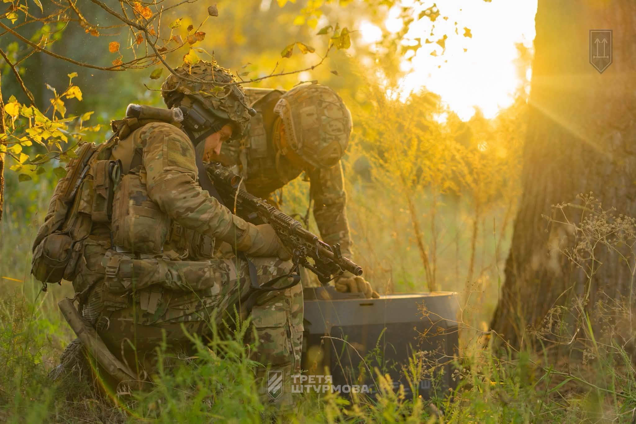 Two soldiers in helmets bend over a box of electronic warfare equipment in the bushes. Sunset in the background.