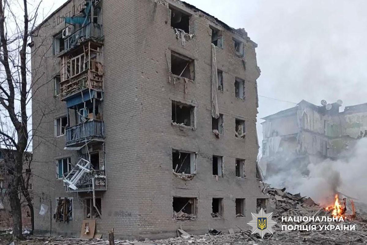 A multi-storey brick building with blown-out windows and wrecked balconies; next to it, burning debris and a collapsed building.
