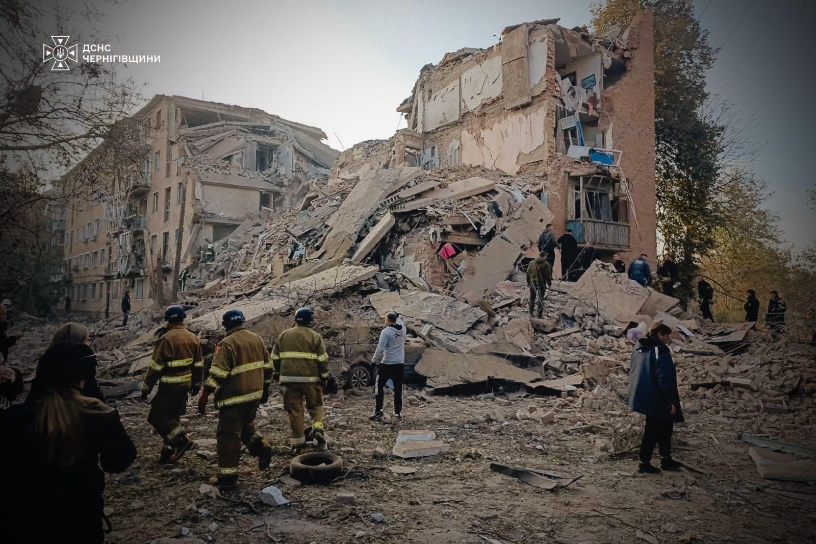 State Emergency Service rescuers in yellow jackets walk across the rubble of a destroyed residential building; residents stand around.
