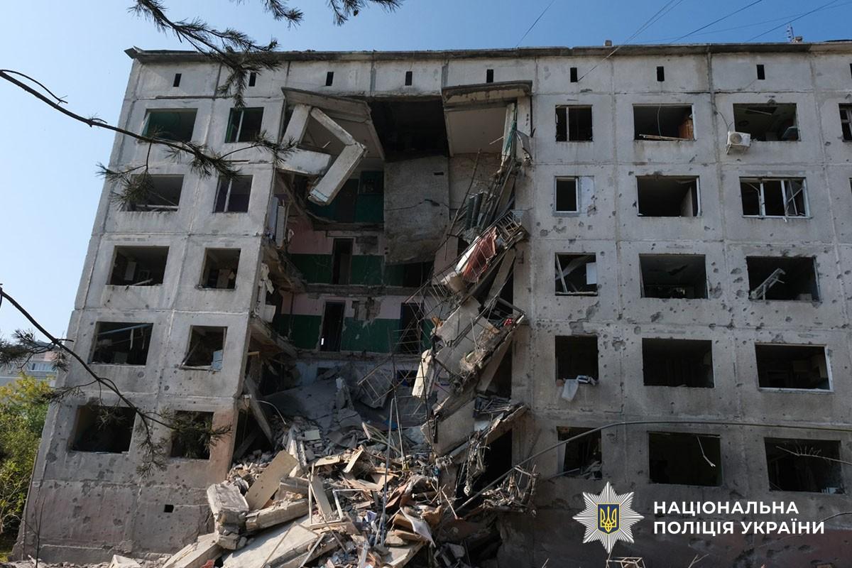 A large hole in the middle of a multi-storey prefabricated block of flats, debris by the wall, blown-out windows and shrapnel marks.