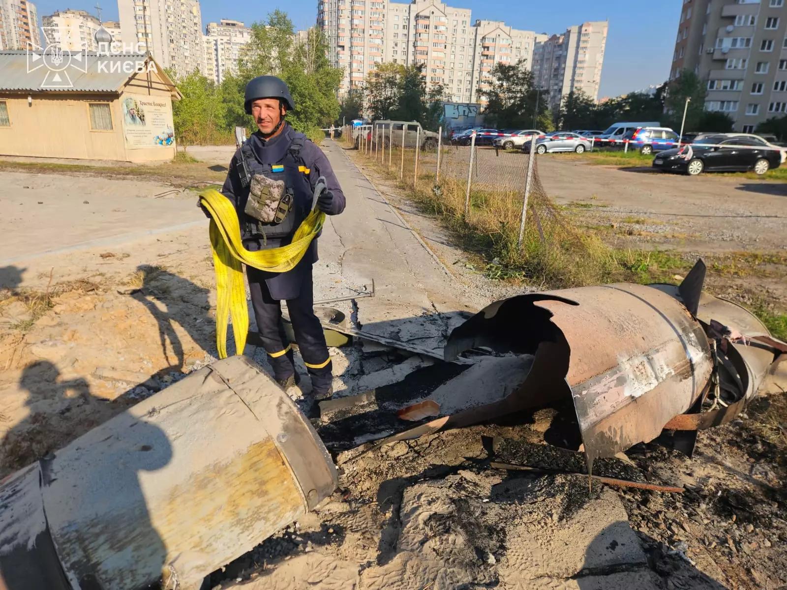 Рятувальник у шоломі й темній формі стоїть поруч із покрученими металевими уламками великої ракети, що лежать на розкопаному піску. На задньому плані – багатоповерхові житлові будинки та дерева.