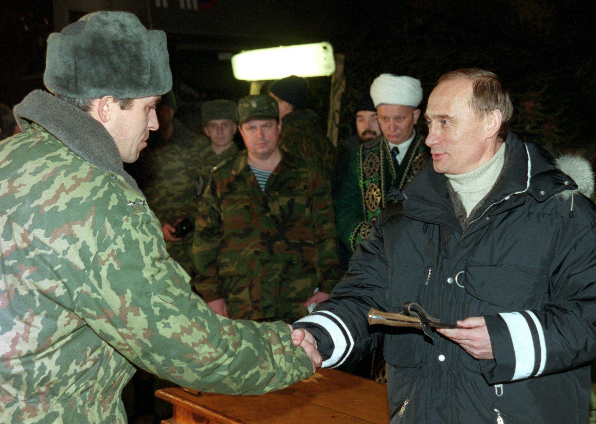 Владімір Путін у темній куртці тисне руку солдатові в хутряній шапці та польовій формі, у другій руці тримає коробку з нагородою. На задньому плані – група військових і чоловік у білій папасі.