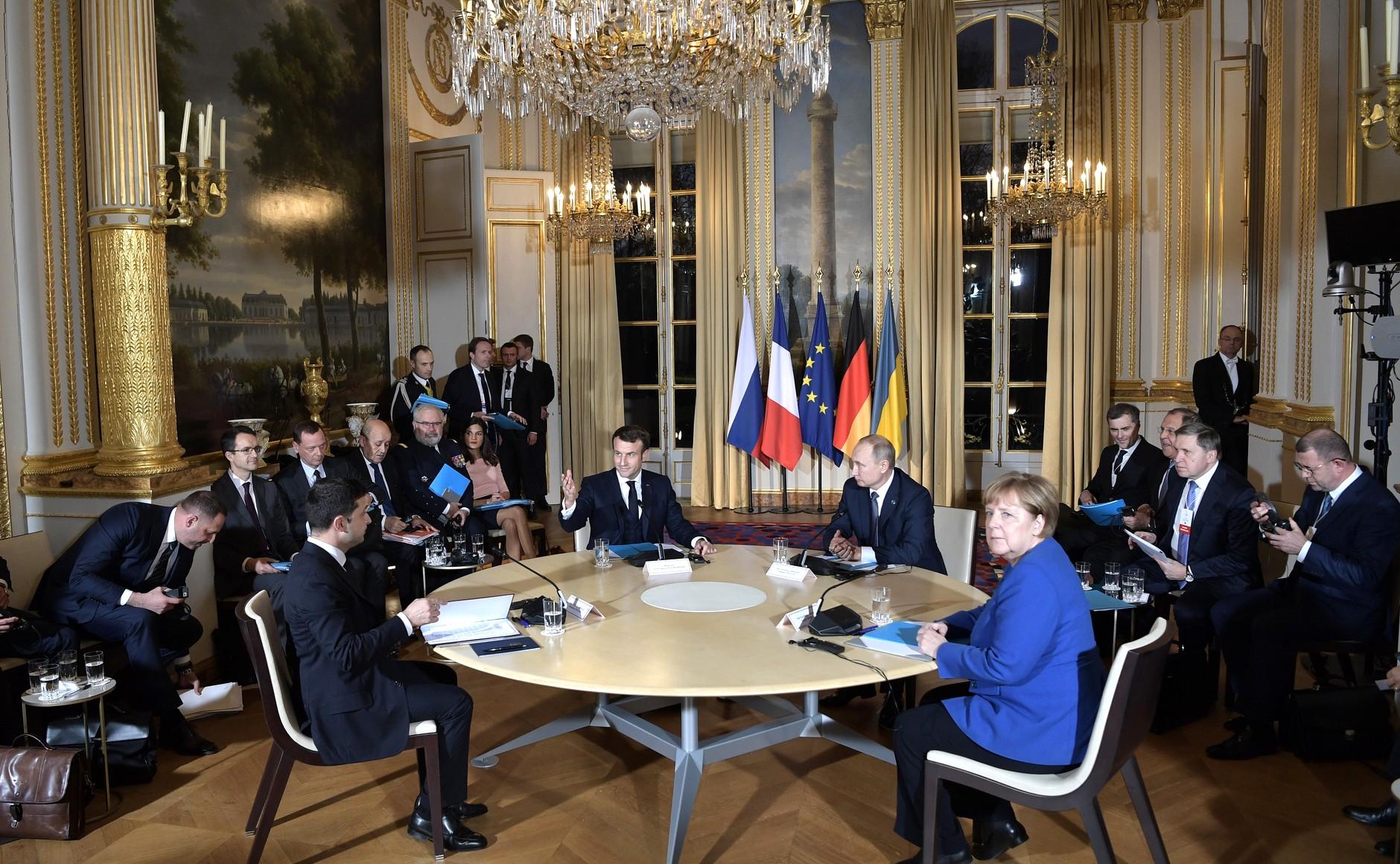 The leaders of France, Germany, Russia and Ukraine with their delegations at a round table in an ornate palace hall with a chandelier and the flags of the four states and the European Union.