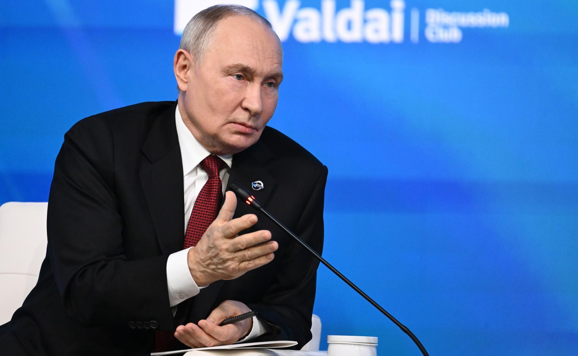 Vladimir Putin sits in a white armchair on stage next to the moderator, gesturing; behind them a blue wall with the Valdai Club logo.
