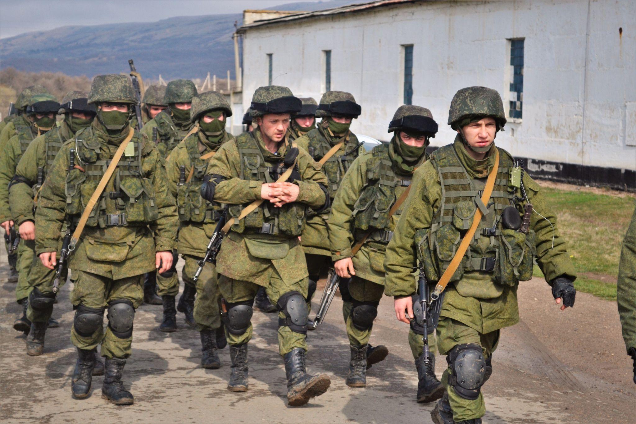 A group of soldiers in helmets and field uniforms without insignia, carrying rifles, walks along a road past buildings of a Ukrainian military base in Crimea.